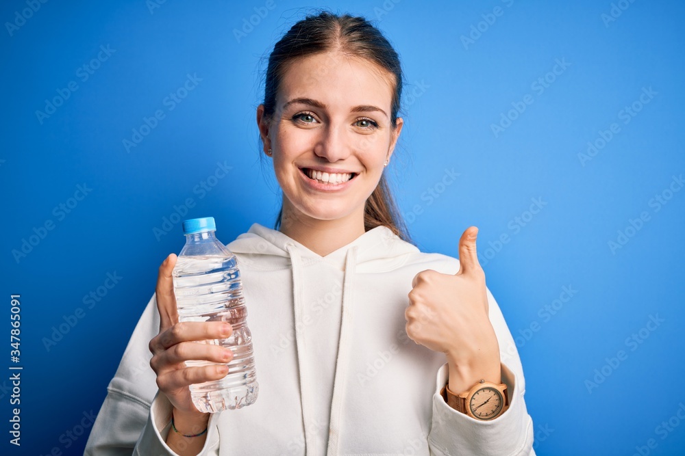 Young beautiful redhead woman doing sport drinking bottle of water over blue background happy with big smile doing ok sign, thumb up with fingers, excellent sign Young beautiful redhead woman doing sport drinking bottle of water over blue background happy with big smile doing ok sign, thumb up with fingers, excellent sign