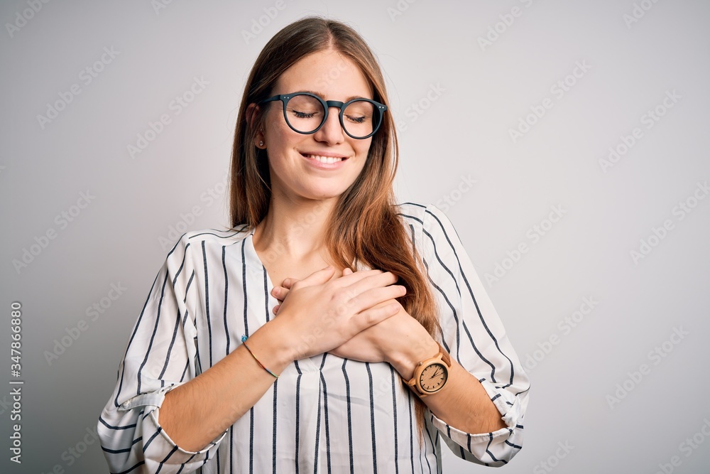 Young beautiful redhead woman wearing casual striped t-shirt and glasses smiling with hands on chest with closed eyes and grateful gesture on face. Health concept.