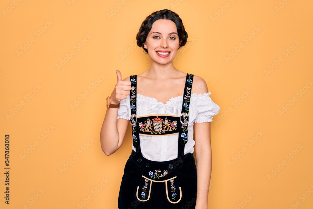 Young beautiful german woman with blue eyes wearing traditional octoberfest dress doing happy thumbs up gesture with hand. Approving expression looking at the camera showing success. Young beautiful german woman with blue eyes wearing traditional octoberfest dress doing happy thumbs up gesture with hand. Approving expression looking at the camera showing success.