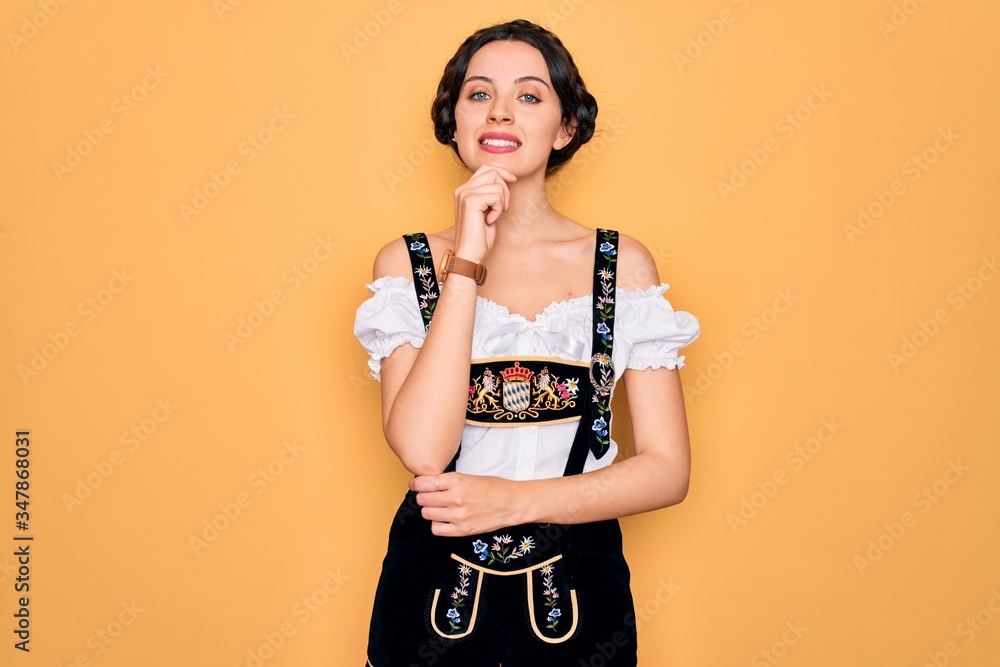 Young beautiful german woman with blue eyes wearing traditional octoberfest dress looking confident at the camera with smile with crossed arms and hand raised on chin. Thinking positive. Young beautiful german woman with blue eyes wearing traditional octoberfest dress looking confident at the camera with smile with crossed arms and hand raised on chin. Thinking positive.