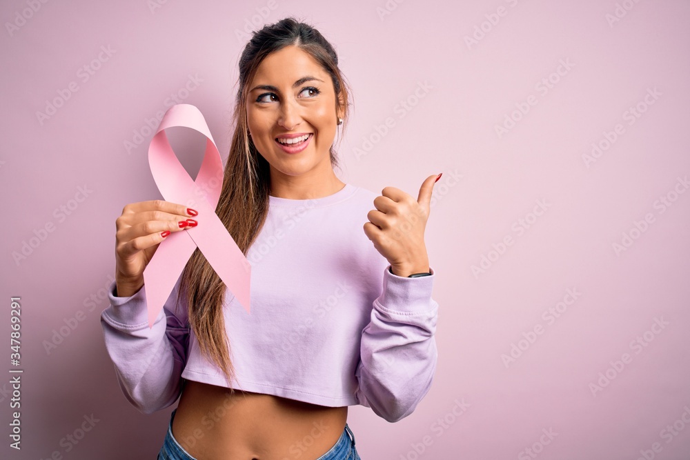 Young beautiful brunette woman holding pink cancer ribbon symbol over isolated background pointing and showing with thumb up to the side with happy face smiling Young beautiful brunette woman holding pink cancer ribbon symbol over isolated background pointing and showing with thumb up to the side with happy face smiling