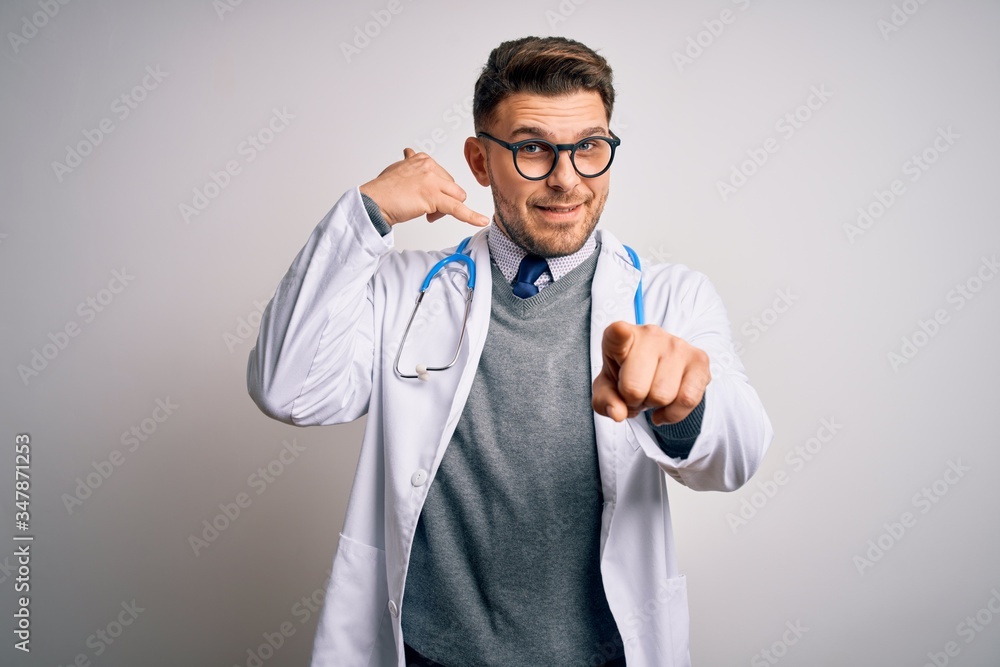 Young doctor man with blue eyes wearing medical coat and stethoscope over isolated background smiling doing talking on the telephone gesture and pointing to you. Call me. Young doctor man with blue eyes wearing medical coat and stethoscope over isolated background smiling doing talking on the telephone gesture and pointing to you. Call me.