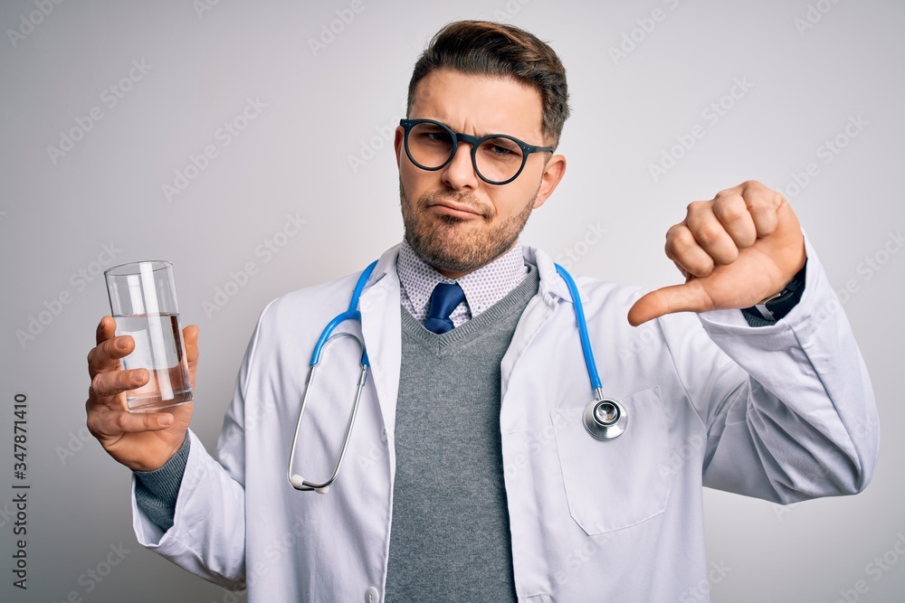 Young doctor man wearing medical coat holding a glass of fresh water over isolated background with angry face, negative sign showing dislike with thumbs down, rejection concept Young doctor man wearing medical coat holding a glass of fresh water over isolated background with angry face, negative sign showing dislike with thumbs down, rejection concept
