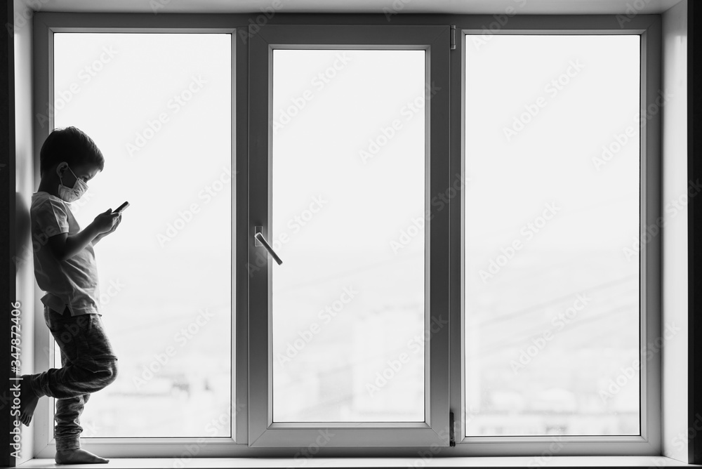 A little boy in a medical mask is sitting on the window of the house in quarantine with a phone in his hands.Prevention of coronavirus and Covid - 19 A little boy in a medical mask is sitting on the window of the house in quarantine with a phone in his hands.Prevention of coronavirus and Covid - 19