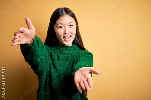 Young beautiful asian woman wearing green winter sweater over yellow isolated background looking at the camera smiling with open arms for hug. Cheerful expression embracing happiness.
