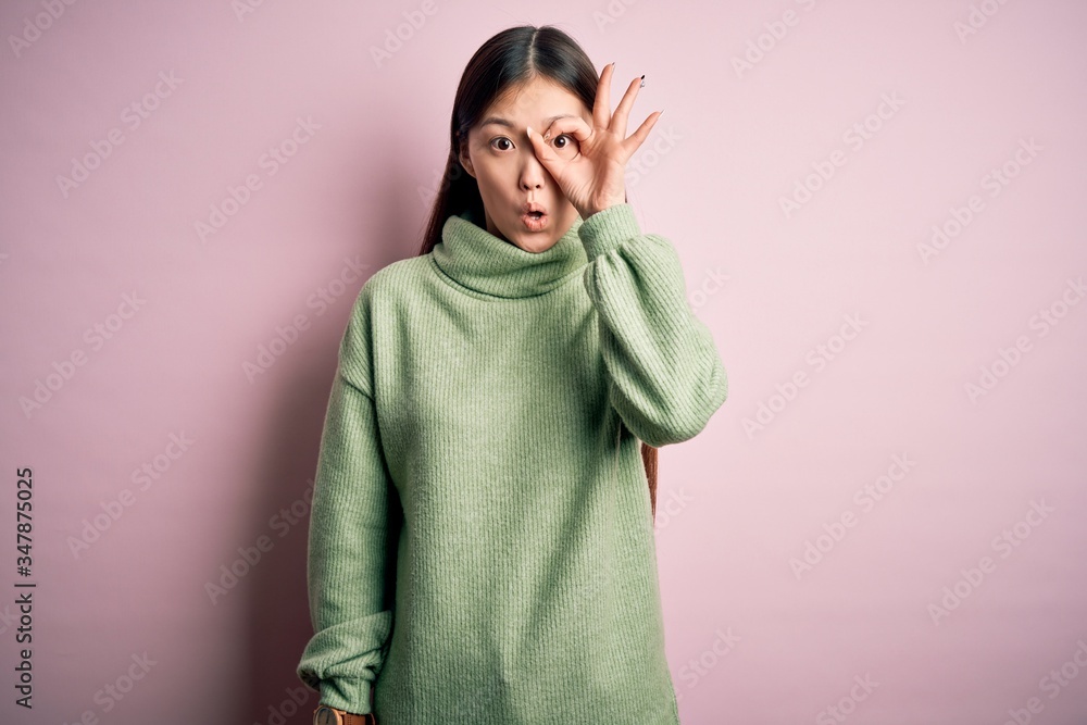 Young beautiful asian woman wearing green winter sweater over pink solated background doing ok gesture shocked with surprised face, eye looking through fingers. Unbelieving expression. Young beautiful asian woman wearing green winter sweater over pink solated background doing ok gesture shocked with surprised face, eye looking through fingers. Unbelieving expression.