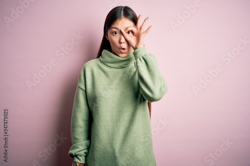 Young beautiful asian woman wearing green winter sweater over pink solated background doing ok gesture shocked with surprised face, eye looking through fingers. Unbelieving expression. © Krakenimages.com