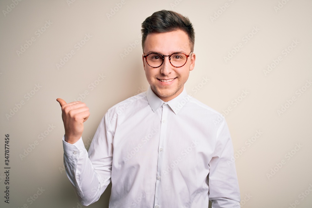Young handsome business mas wearing glasses and elegant shirt over isolated background smiling with happy face looking and pointing to the side with thumb up. Young handsome business mas wearing glasses and elegant shirt over isolated background smiling with happy face looking and pointing to the side with thumb up.