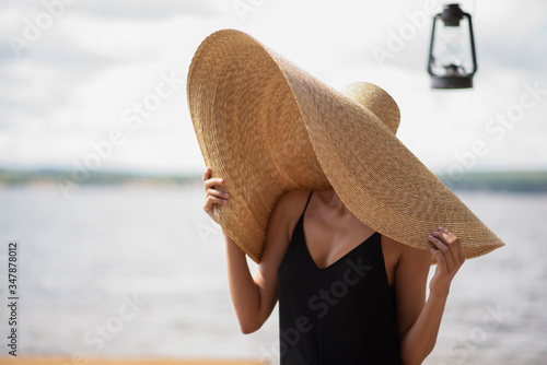A young woman on a beach wearing a giant straw hat and a lighting is close to her 