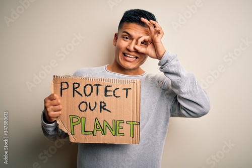 Young handsome activist latin man holding banner asking to protect our planet with happy face smiling doing ok sign with hand on eye looking through fingers © Krakenimages.com