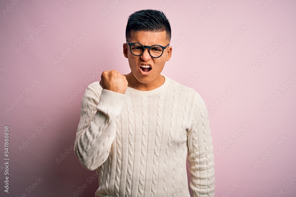 Young handsome latin man wearing white casual sweater and glasses over pink background angry and mad raising fist frustrated and furious while shouting with anger. Rage and aggressive concept. Young handsome latin man wearing white casual sweater and glasses over pink background angry and mad raising fist frustrated and furious while shouting with anger. Rage and aggressive concept.