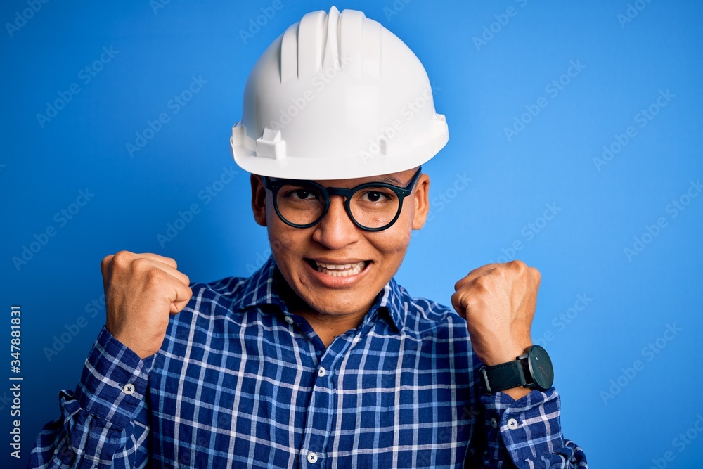 Young handsome engineer latin man wearing safety helmet over isolated blue background celebrating surprised and amazed for success with arms raised and open eyes. Winner concept.