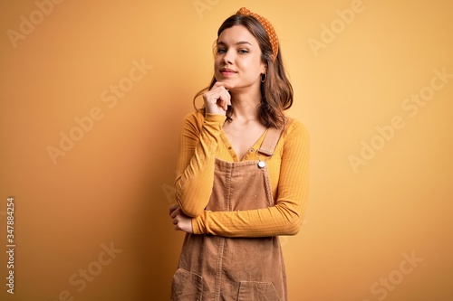 Young beautiful blonde girl wearing overall standing over yellow isolated background looking confident at the camera smiling with crossed arms and hand raised on chin. Thinking positive.