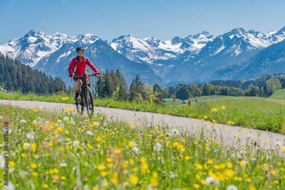 pretty senior woman riding her electric mountain on warm spring day with snoe capped bike in the Iller valley near Oberstdorf with snoe capped Allgau High Alps in background, Allgau Alps, Bavaria, 