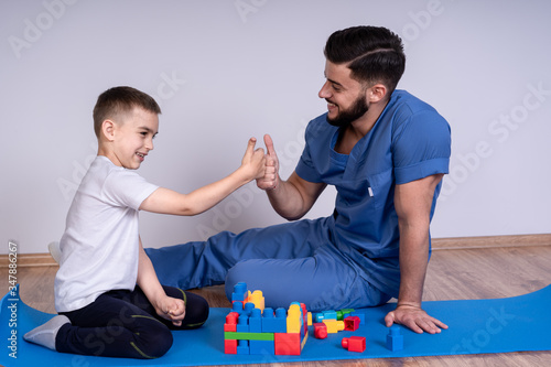 Young male doctor with beard in a blue uniform sitting on the floor next to the boy 10 years, they play educational toys