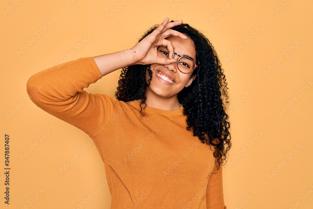 Young african american woman wearing casual sweater and glasses over yellow background doing ok gesture with hand smiling, eye looking through fingers with happy face. Young african american woman wearing casual sweater and glasses over yellow background doing ok gesture with hand smiling, eye looking through fingers with happy face.
