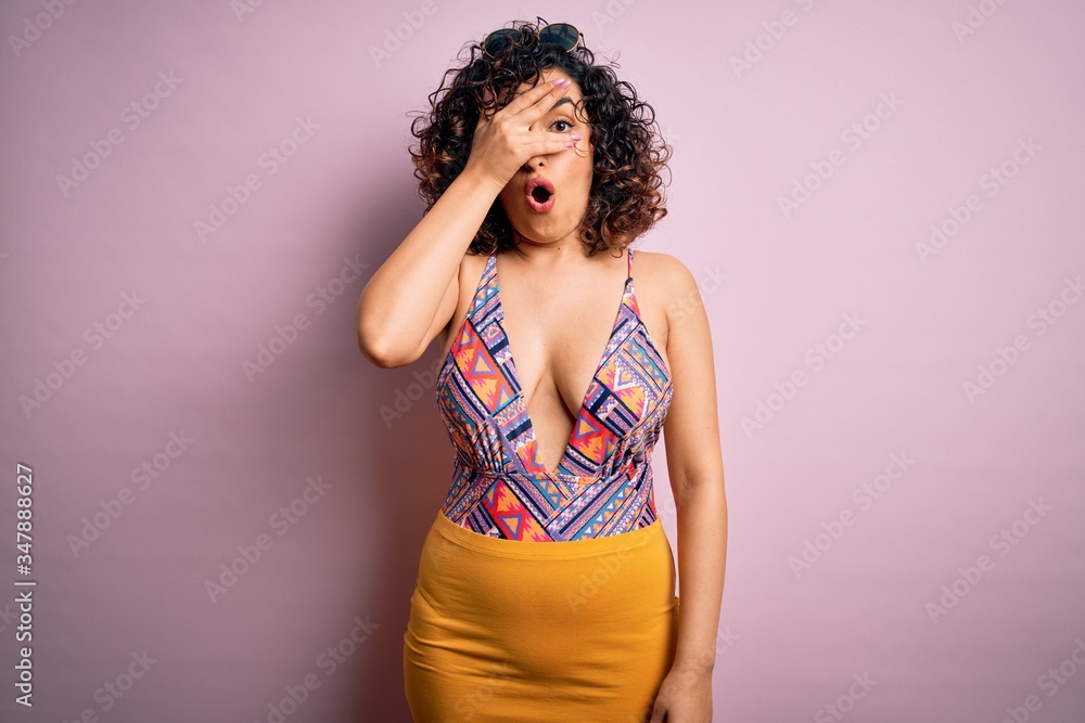 Young beautiful arab woman on vacation wearing swimsuit and sunglasses over pink background peeking in shock covering face and eyes with hand, looking through fingers with embarrassed expression. Young beautiful arab woman on vacation wearing swimsuit and sunglasses over pink background peeking in shock covering face and eyes with hand, looking through fingers with embarrassed expression.