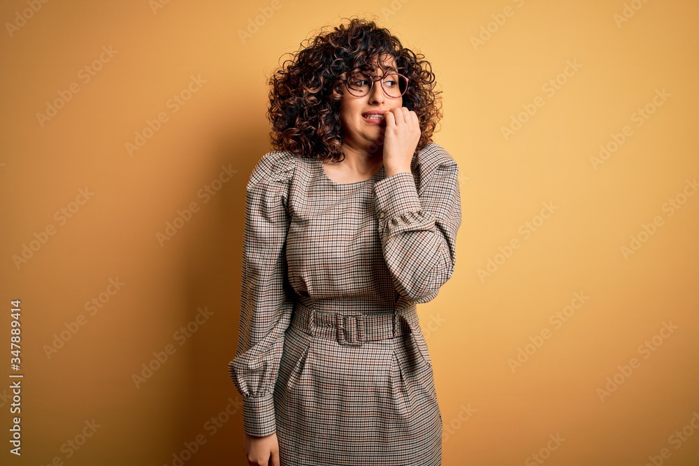 Beautiful arab business woman wearing dress and glasses standing over yellow background looking stressed and nervous with hands on mouth biting nails. Anxiety problem. Beautiful arab business woman wearing dress and glasses standing over yellow background looking stressed and nervous with hands on mouth biting nails. Anxiety problem.