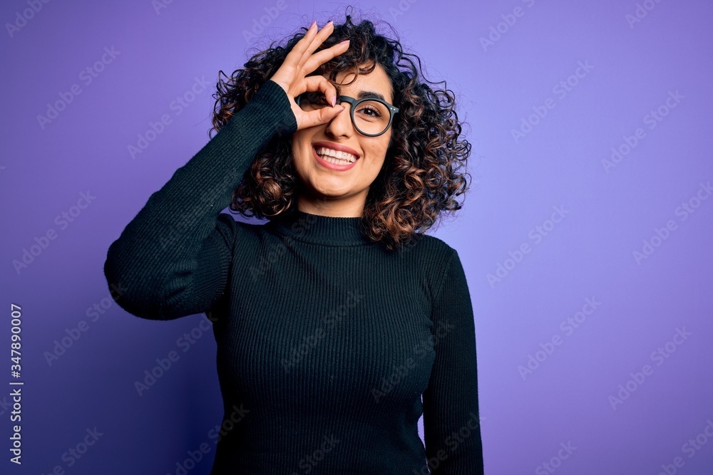 Young beautiful curly arab woman wearing casual sweater and glasses over purple background doing ok gesture with hand smiling, eye looking through fingers with happy face. Young beautiful curly arab woman wearing casual sweater and glasses over purple background doing ok gesture with hand smiling, eye looking through fingers with happy face.