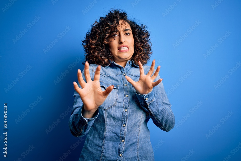 Young beautiful curly arab woman wearing casual denim shirt standing over blue background disgusted expression, displeased and fearful doing disgust face because aversion reaction. With hands raised Young beautiful curly arab woman wearing casual denim shirt standing over blue background disgusted expression, displeased and fearful doing disgust face because aversion reaction. With hands raised