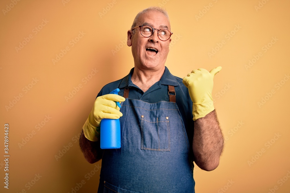 Middle age hoary cleaner man cleaning wearing apron and gloves using sprayer pointing and showing with thumb up to the side with happy face smiling Middle age hoary cleaner man cleaning wearing apron and gloves using sprayer pointing and showing with thumb up to the side with happy face smiling