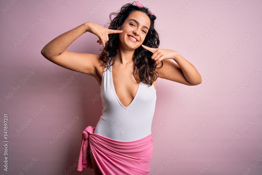 Beautiful woman with curly hair on vacation wearing white swimsuit over pink background smiling cheerful showing and pointing with fingers teeth and mouth. Dental health concept. Beautiful woman with curly hair on vacation wearing white swimsuit over pink background smiling cheerful showing and pointing with fingers teeth and mouth. Dental health concept.