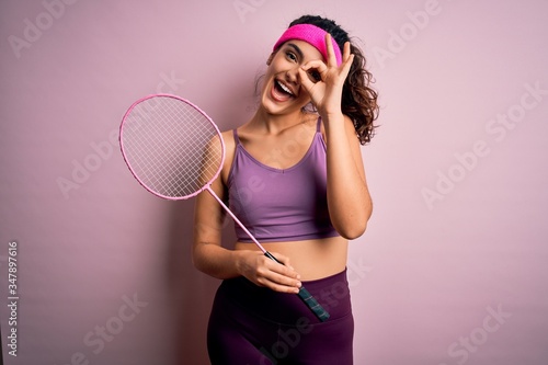 Beautiful sportswoman with curly hair wearing sportswear playing badminton uning racket with happy face smiling doing ok sign with hand on eye looking through fingers © Krakenimages.com