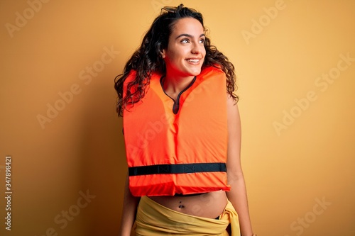 Young beautiful woman with curly hair wearing orange lifejacket over yellow background looking away to side with smile on face, natural expression. Laughing confident.