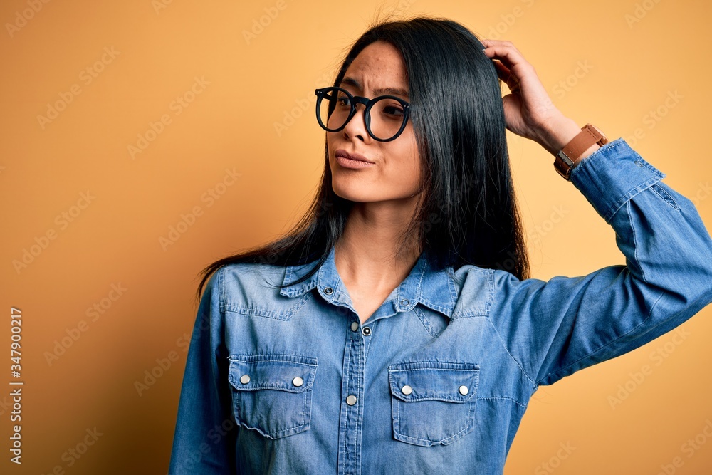 Young beautiful chinese woman wearing casual denim shirt over isolated yellow background confuse and wondering about question. Uncertain with doubt, thinking with hand on head. Pensive concept.