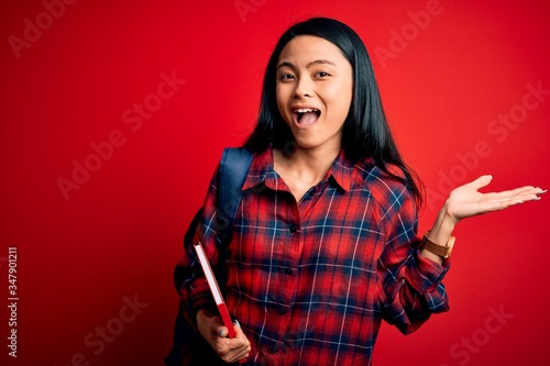Young beautiful chinese student woman holding book standing over isolated pink background very happy and excited, winner expression celebrating victory screaming with big smile and raised hands