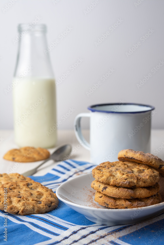 Close-up of chocolate chip cookies on white plate with cup, spoon and bottle of milk on blue napkin, with selective focus, white background, vertical Close-up of chocolate chip cookies on white plate with cup, spoon and bottle of milk on blue napkin, with selective focus, white background, vertical