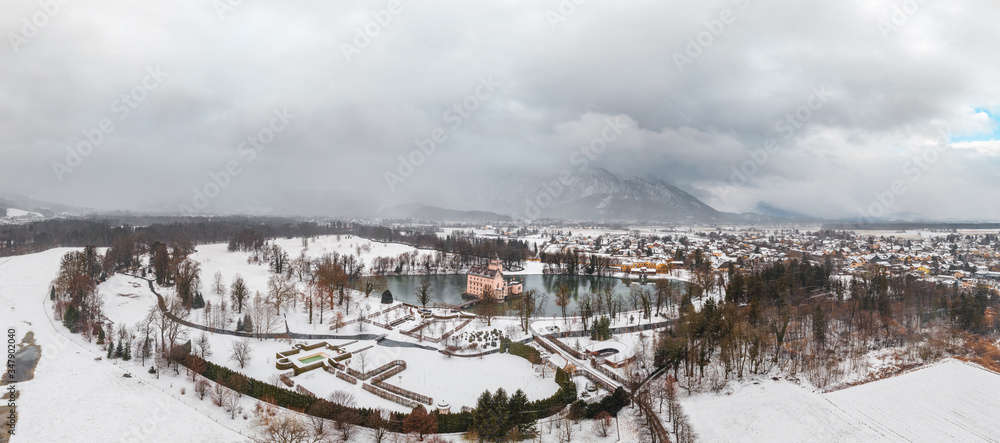 Panoramic aerial view of Schloss Anif castle moated in artificial pond near Salzburg outskirts in heavy snow during winter with view of untersberg