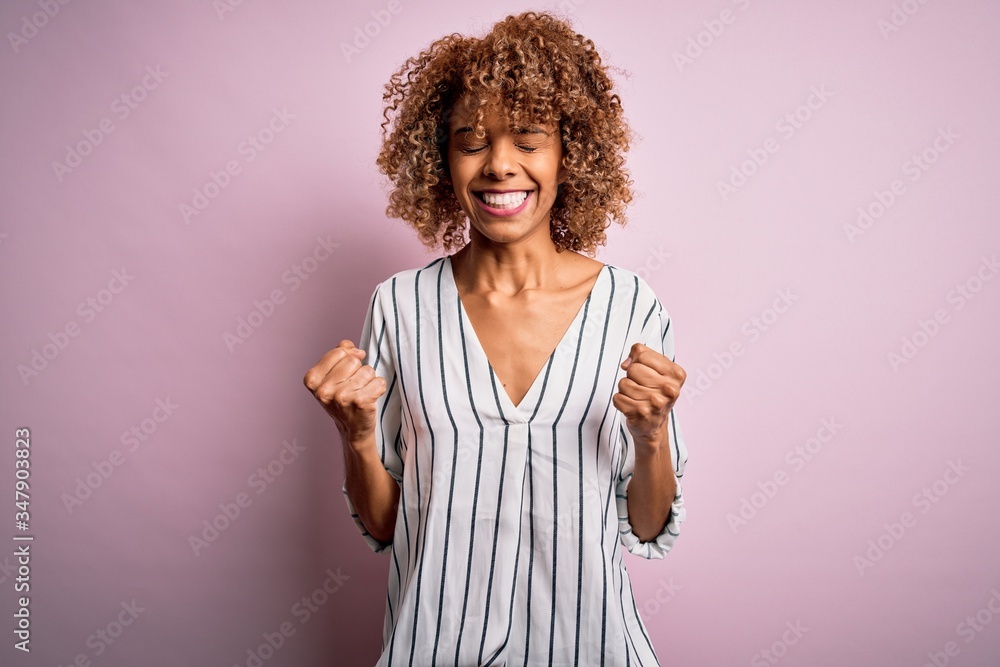 Beautiful african american woman with curly hair wearing striped t-shirt over pink background excited for success with arms raised and eyes closed celebrating victory smiling. Winner concept. Beautiful african american woman with curly hair wearing striped t-shirt over pink background excited for success with arms raised and eyes closed celebrating victory smiling. Winner concept.