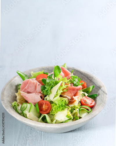 Salad with ham, vine tomatoes, parmesan cheese, prosciutto and green mix over light blue background with copy space. Close up. Healthy food concept.