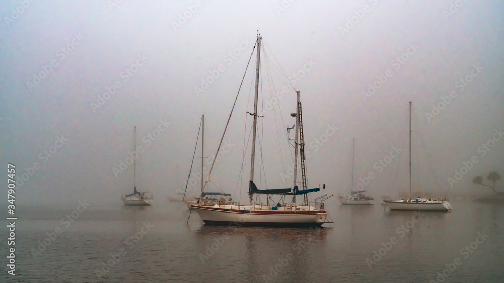Sailboats anchored in thick grey foggy harbor in the morning