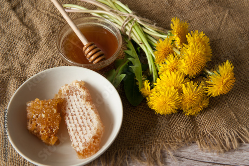 A bouquet of yellow dandelion flowers and a Cup with honey and honeycomb on a burlap on a wooden background. Alternative medicine, medicinal herbs. Selective focus.