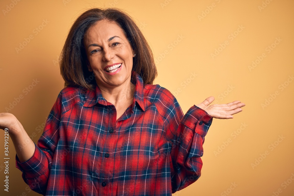 Middle age beautiful woman wearing casual shirt standing over isolated yellow background smiling showing both hands open palms, presenting and advertising comparison and balance Middle age beautiful woman wearing casual shirt standing over isolated yellow background smiling showing both hands open palms, presenting and advertising comparison and balance