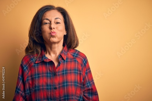 Middle age beautiful woman wearing casual shirt standing over isolated yellow background puffing cheeks with funny face. Mouth inflated with air, crazy expression.