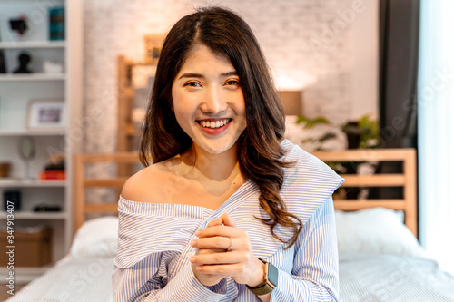 Portrait of smiling young Asian girl sitting on bed while making a video call in bedroom at home. Video conferencing technology and wireless communication concept - Headshot point of view.