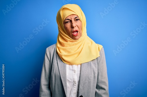 Middle age brunette business woman wearing muslim traditional hijab over blue background In shock face, looking skeptical and sarcastic, surprised with open mouth © Krakenimages.com