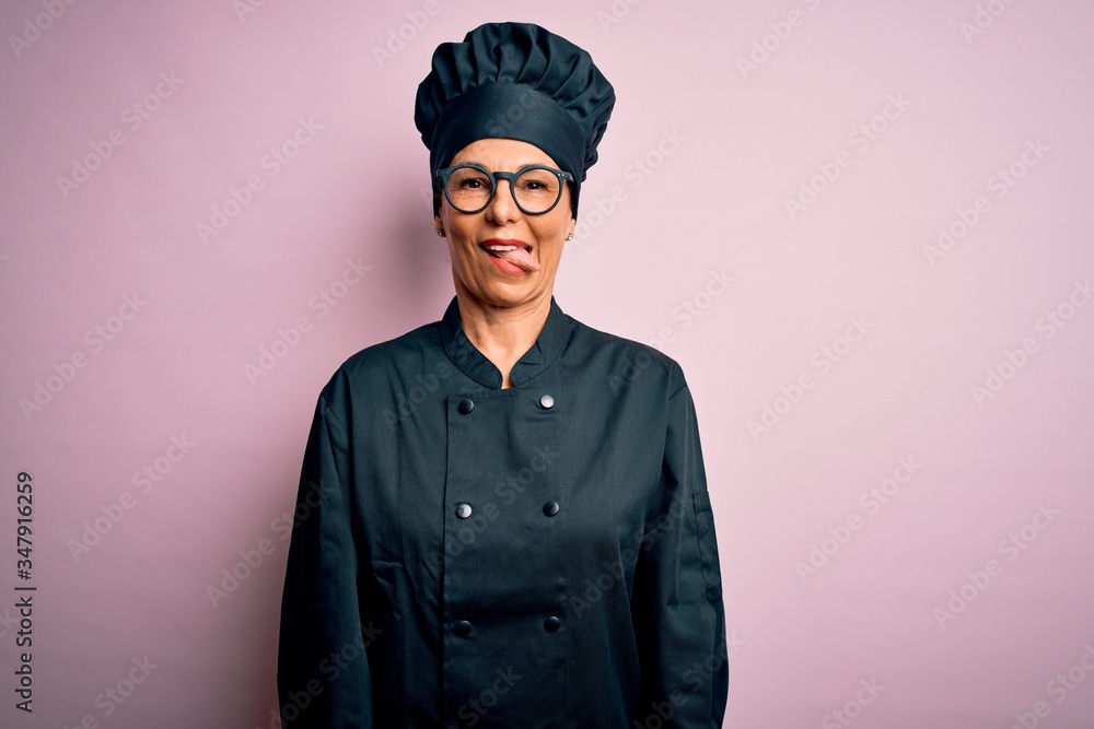 Middle age brunette chef woman wearing cooker uniform and hat over isolated pink background sticking tongue out happy with funny expression. Emotion concept. Middle age brunette chef woman wearing cooker uniform and hat over isolated pink background sticking tongue out happy with funny expression. Emotion concept.