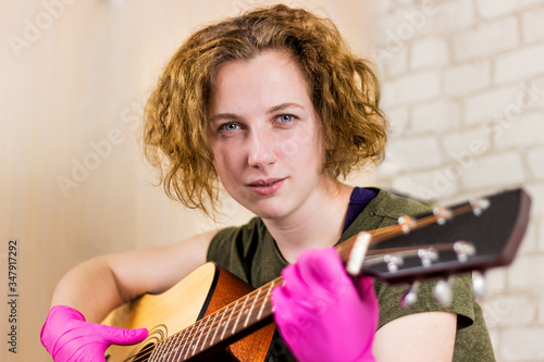 A young pretty teen girl stares at the camera and plays the guitar in rubber gloves, learning chords or a song. Getting out of quarantine. Fastidiousness. Misophobia