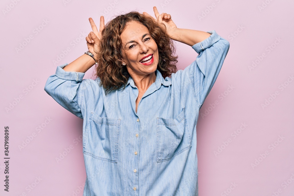 Middle age beautiful woman wearing casual denim shirt standing over pink background Posing funny and crazy with fingers on head as bunny ears, smiling cheerful Middle age beautiful woman wearing casual denim shirt standing over pink background Posing funny and crazy with fingers on head as bunny ears, smiling cheerful