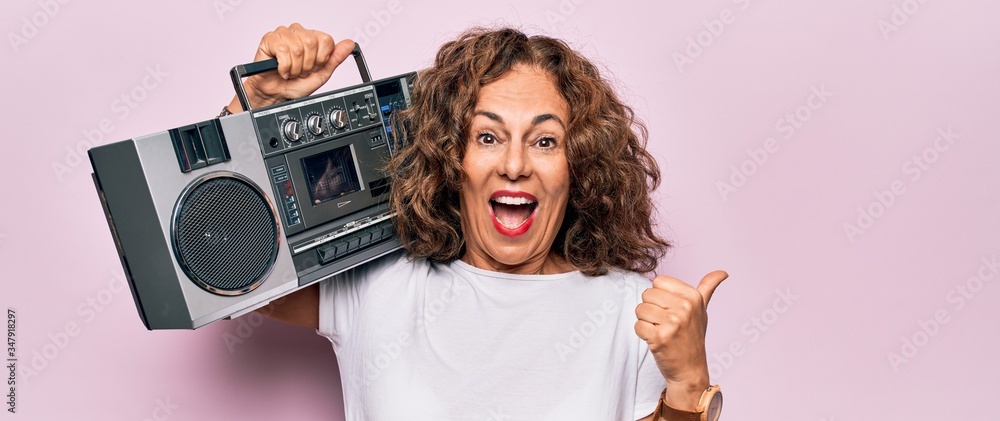 Middle age brunette hipster woman holding retro music boombox over pink isolated background pointing thumb up to the side smiling happy with open mouth