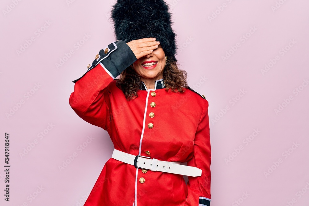 Middle age beautiful wales guard woman wearing traditional uniform over pink background smiling and laughing with hand on face covering eyes for surprise. Blind concept.