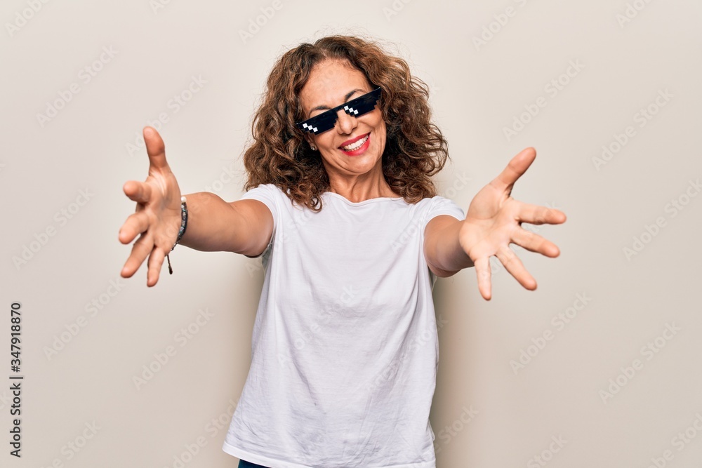 Middle age beautiful woman wearing funny thug life sunglasses over white background looking at the camera smiling with open arms for hug. Cheerful expression embracing happiness. Middle age beautiful woman wearing funny thug life sunglasses over white background looking at the camera smiling with open arms for hug. Cheerful expression embracing happiness.