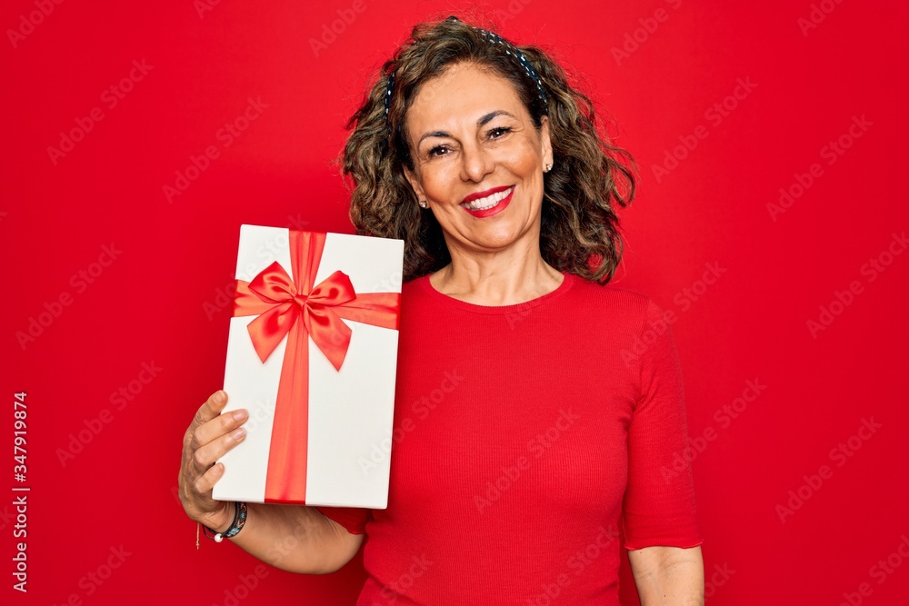 Middle age senior woman holding romantic present box over red isolated background with a happy face standing and smiling with a confident smile showing teeth Middle age senior woman holding romantic present box over red isolated background with a happy face standing and smiling with a confident smile showing teeth