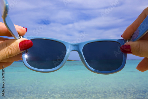 Blue sunglasses in female hands with red manicure against beautiful seascape - clean turqouise water and island on background. Concept of summer travelling, vacation, looking through glasses.