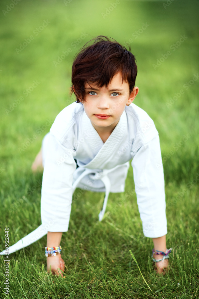 Vertical picture of short haired 7 years old girl in white kimono making push ups karate training outdoors. Martial arts. Sport and activity for children.