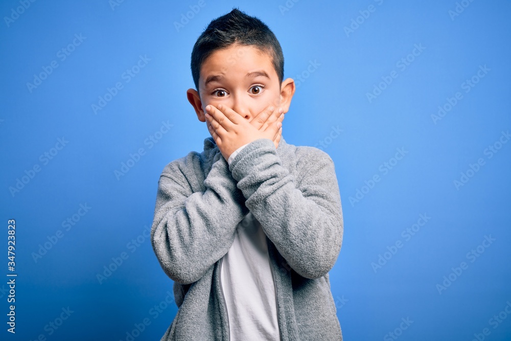 Young little boy kid wearing sport sweatshirt over blue isolated background shocked covering mouth with hands for mistake. Secret concept. Young little boy kid wearing sport sweatshirt over blue isolated background shocked covering mouth with hands for mistake. Secret concept.